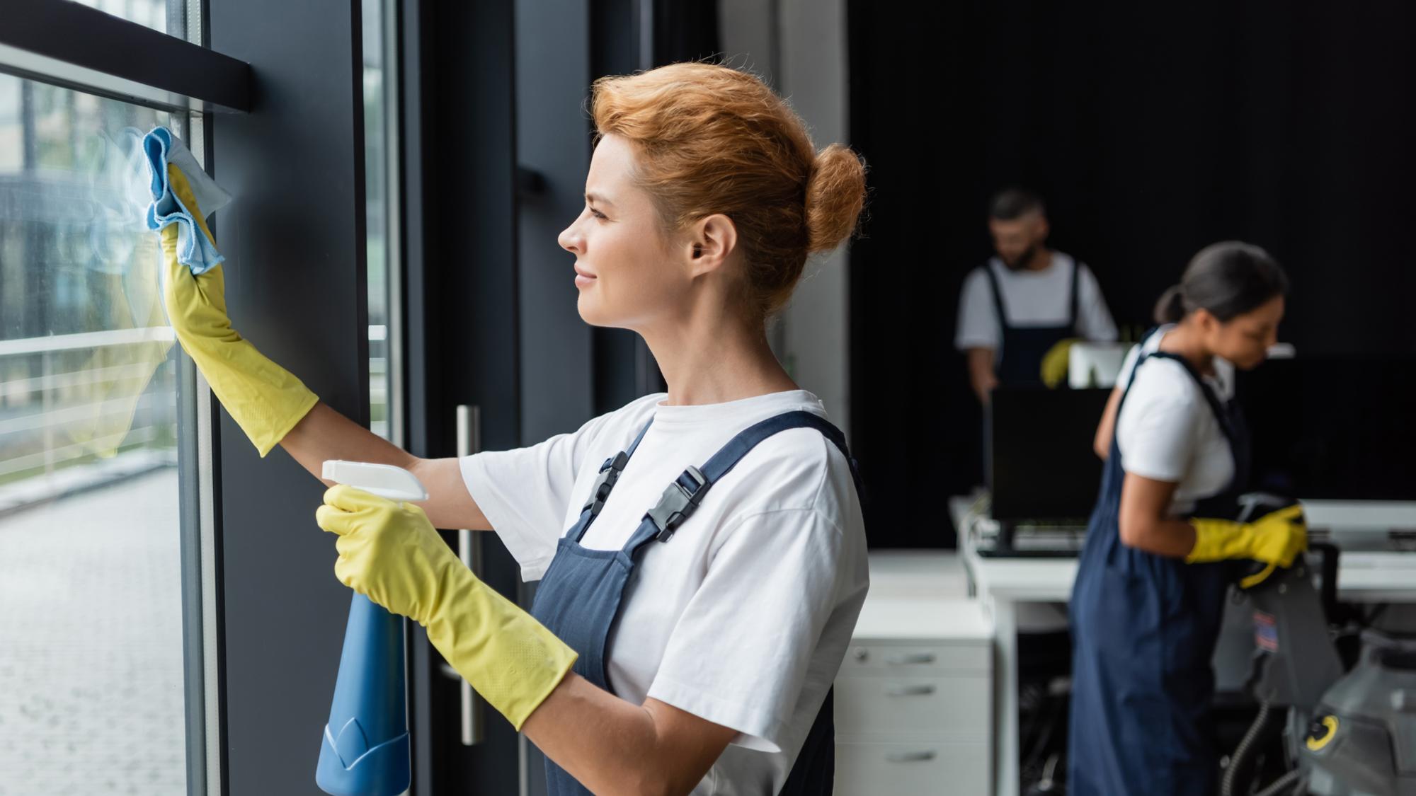 A woman in yellow gloves is cleaning a large glass window with a cloth, smiling as she works. In the background, two people are engaged in cleaning tasks within a modern office space. One person is using a vacuum cleaner, while the other is at a desk. The setting has large windows and a dark backdrop.